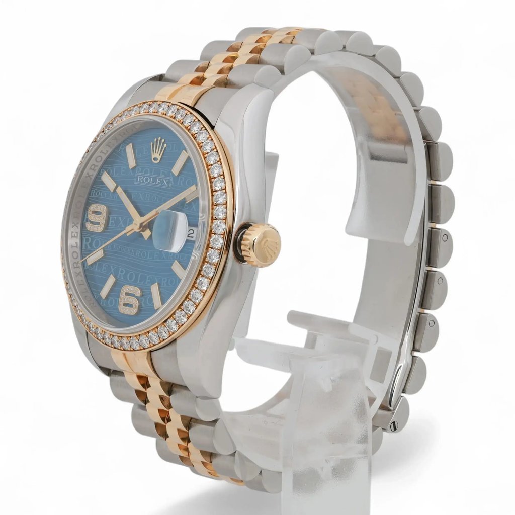 An image of a pre-owned unisex Rolex wristwatch with a two-tone metal bracelet, featuring a prominent blue dial and a diamond-studded bezel. The watch is positioned upright on a transparent stand, viewed from a three-quarter angle, highlighting its date indicator and crown on the right side. The photograph is taken at a close distance, showcasing the intricate details and luxurious design elements.