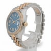 An image of a pre-owned unisex Rolex wristwatch with a two-tone metal bracelet, featuring a prominent blue dial and a diamond-studded bezel. The watch is positioned upright on a transparent stand, viewed from a three-quarter angle, highlighting its date indicator and crown on the right side. The photograph is taken at a close distance, showcasing the intricate details and luxurious design elements.
