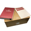 An image of a wristwatch's packaging and documentation, viewed from a slightly elevated angle. The wooden box is centered, with the brand's logo visible on top. To the left, a red operating instructions booklet is placed at an angle. On the right, a white cardholder containing multiple cards is partially visible. The image captures the items at close range, emphasizing the new condition of the packaging materials.