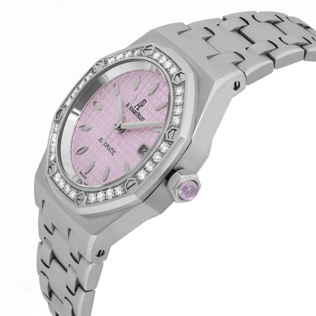 An image of a pre-owned Audemars Piguet Lady Royal Oak 33mm women's wristwatch taken from a slight side angle, showcasing the watch's elegant steel bracelet and pink textured dial. The watch face is surrounded by a diamond-studded bezel, and a round gemstone is set into the crown on the right side. The photograph is taken at a moderate distance, providing a clear view of the watch's intricate details and design.