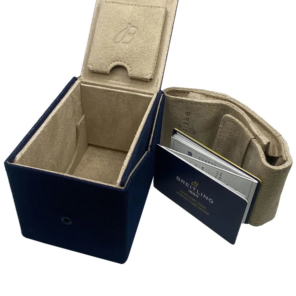 An image of a Breitling men's wristwatch packaging, without the watch present. The view is from a slightly elevated angle, showing the full open box and documents. The box is navy blue with a beige interior, and the Breitling logo is embossed on the lid's interior. The box is positioned to the left and angled to show the interior clearly. To the right, there is a beige cushion for the watch, and a warranty card holder with documents partially visible, both bearing the Breitling logo. The items are in the foreground and occupy most of the frame, suggesting a close-up perspective. 