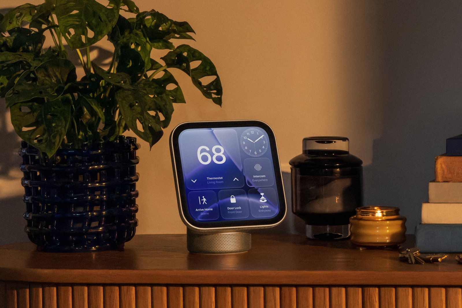 Apple HomeView als Tischgerät auf einer Holzkommode im Wohnzimmer, daneben eine Zimmerpflanze, eine Kerze und Bücher; auf dem Display Thermostat, Uhr und Kacheln für Licht und Türschloss