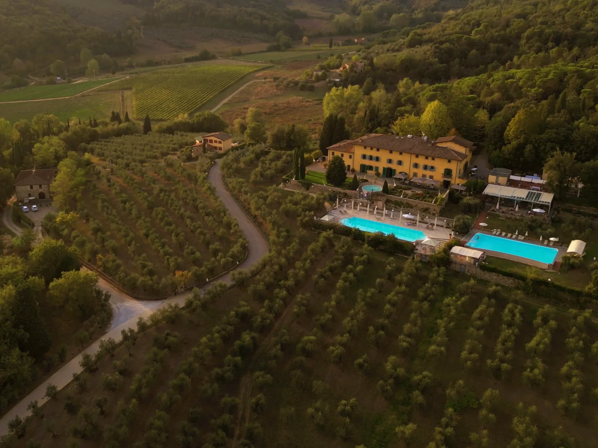 Villa La Palagina, Figline Valdarno: the ochre villa and its two pools seen from the air, among olive groves and vineyards at dusk