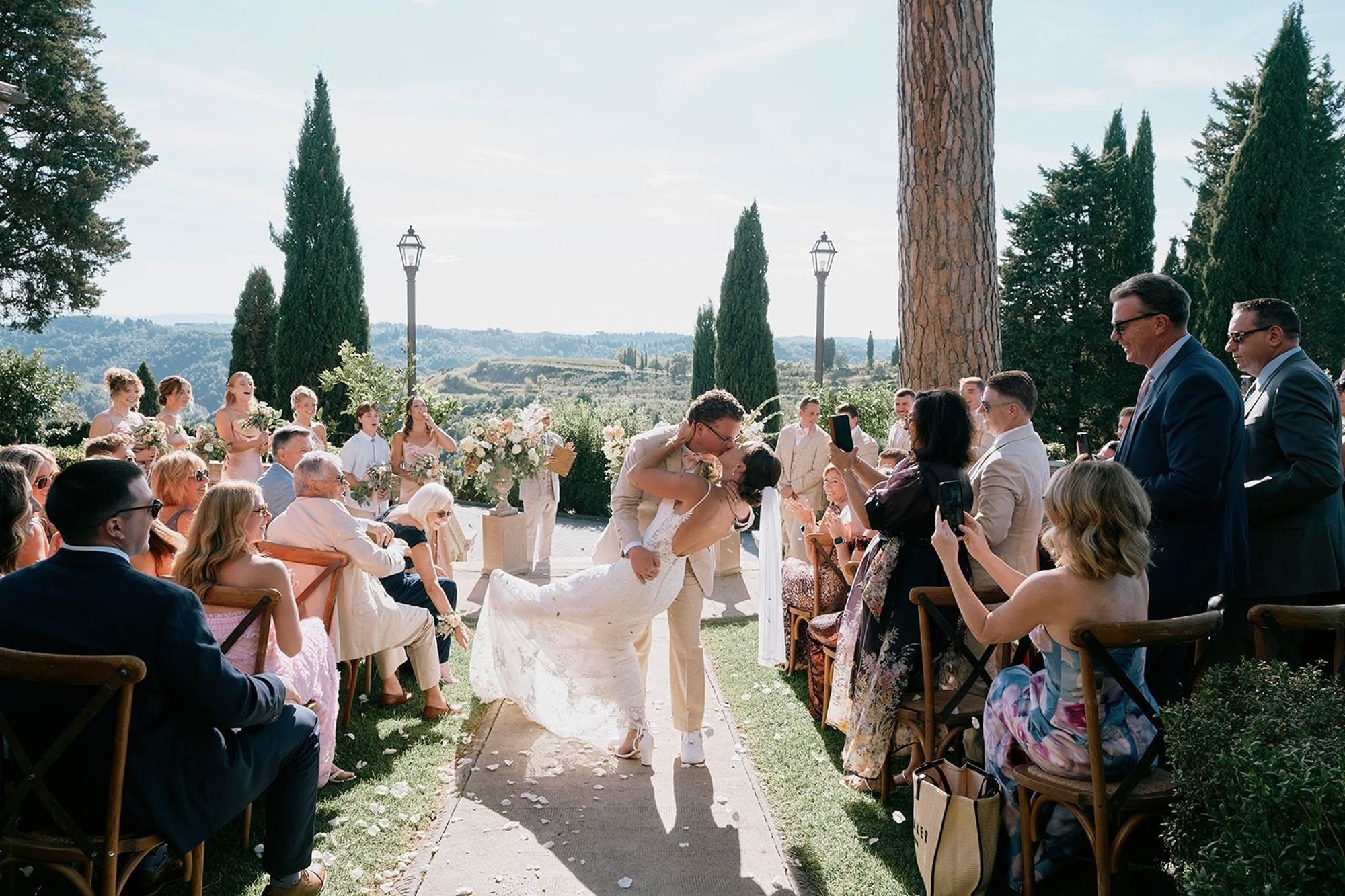 Borgo Bucciano, San Miniato: guests on wooden chairs either side of the aisle as the couple kiss under the cypresses and an umbrella pine