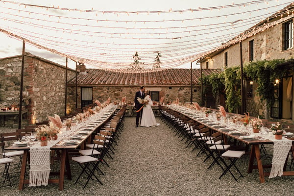 Locanda In Tuscany, Castiglion Fiorentino — two long imperial tables laid for dinner in the stone courtyard, under a canopy of fairy lights at dusk