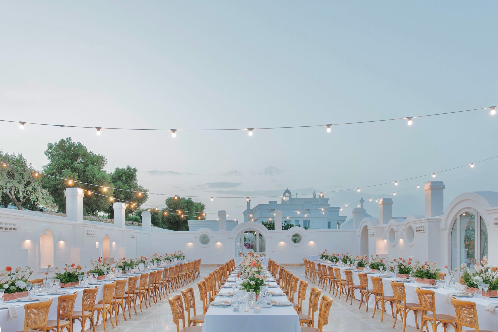 Masseria Borgo Ritella, Fasano: long tables with orange-flowered centrepieces in a white courtyard under festoon lights at dusk