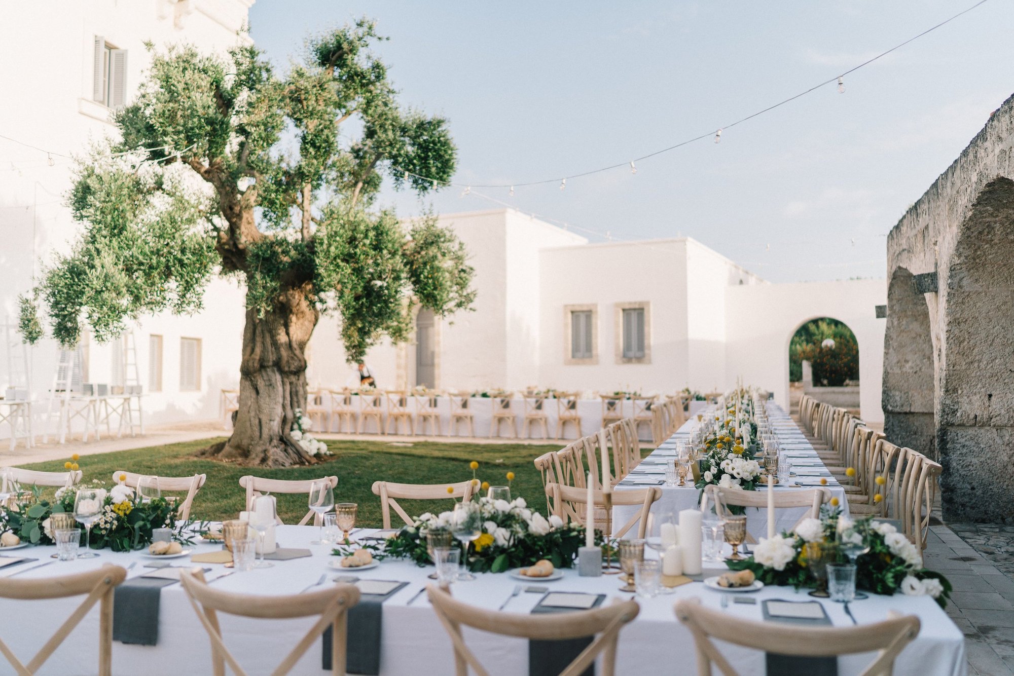 Masseria San Giovanni, Fasano: long tables with white flowers in the whitewashed courtyard, an old olive tree on the lawn beside them