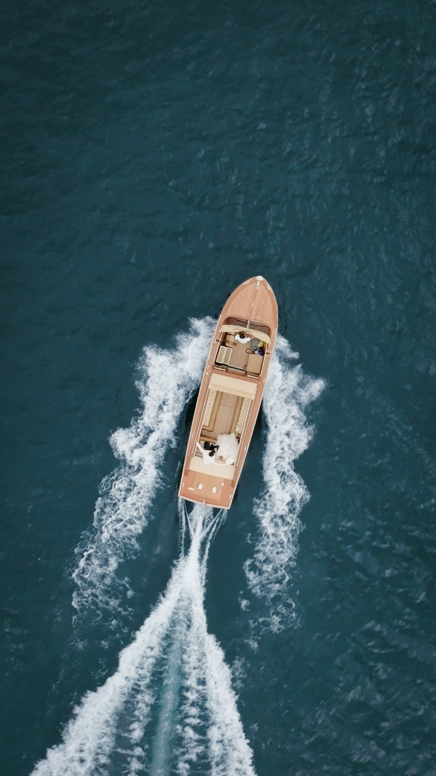 Andrea Tricarico Films — a bride at the stern of a wooden motorboat on Lake Como, filmed from directly above with the wake curling behind her