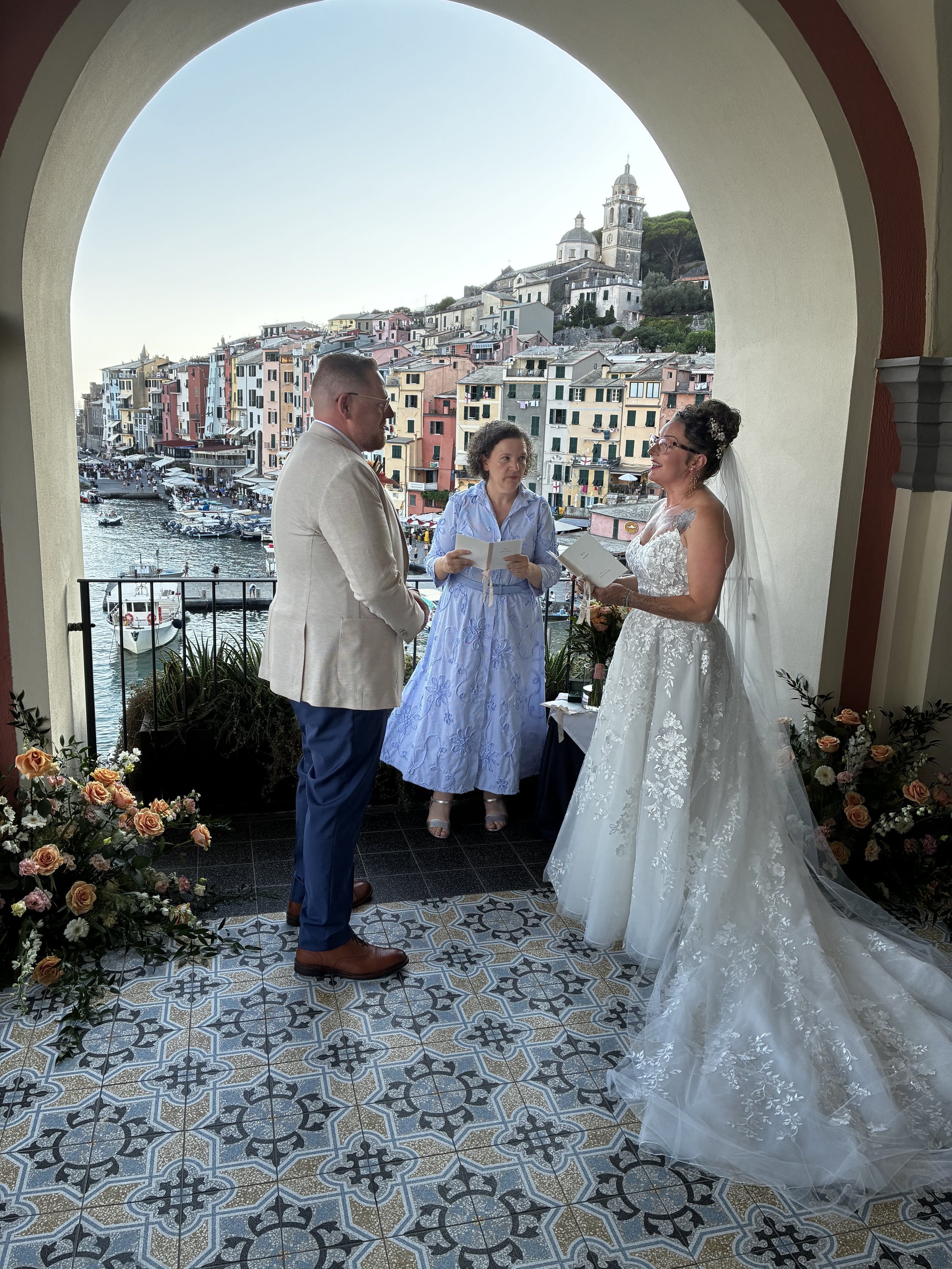 Love Celebrant Italy: a celebrant leading a ceremony for a bride and groom under a stone arch on a tiled terrace, a harbour town of painted houses behind them