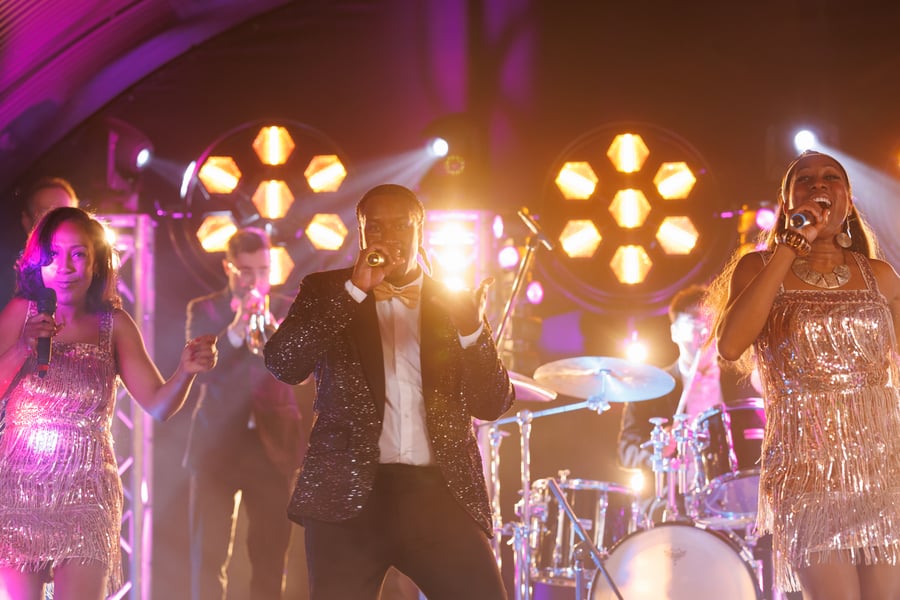Rocks The Room mid-set — the frontman on trumpet with two vocalists in sequinned fringe either side, hex stage lights behind them