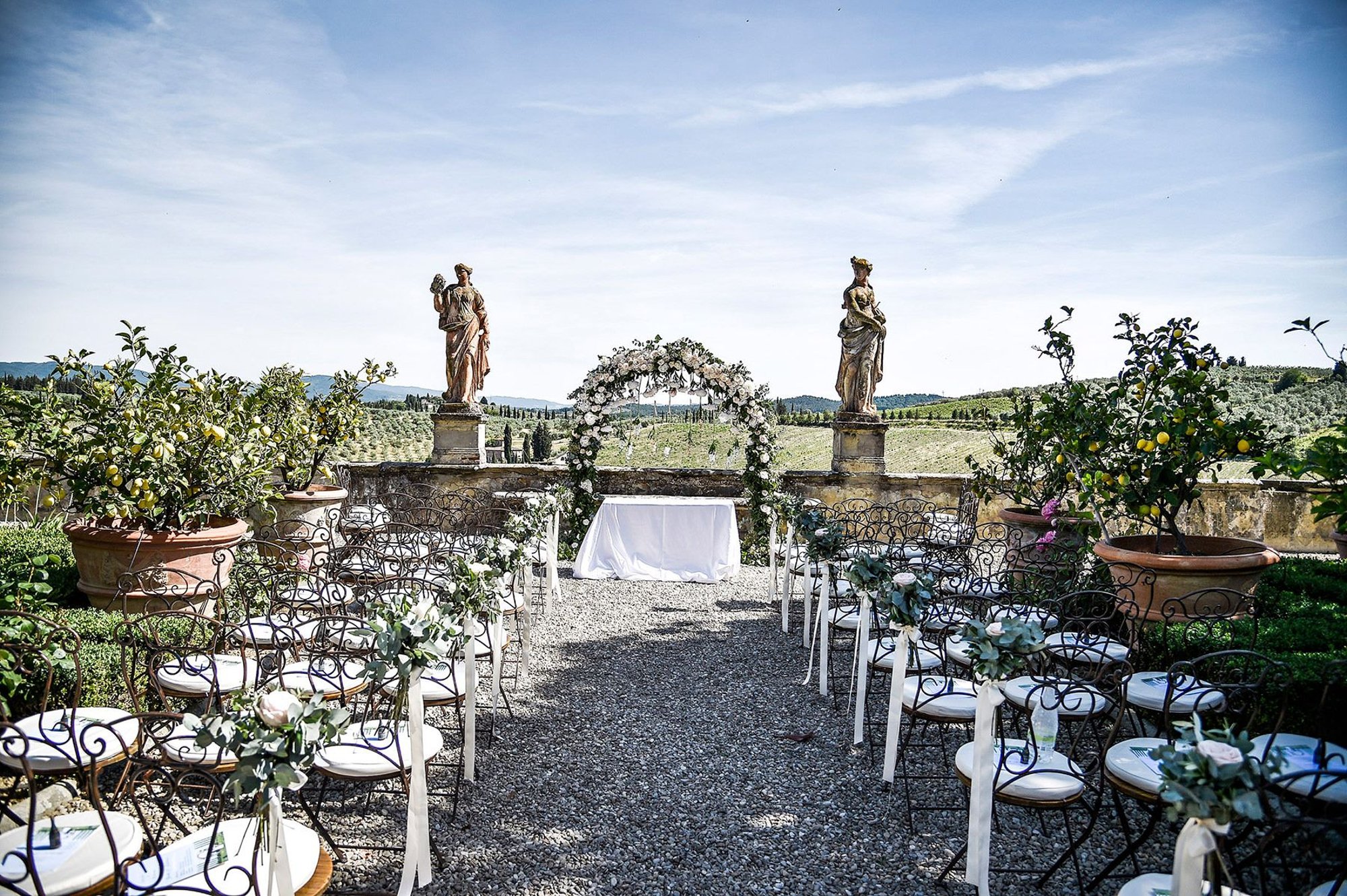 Villa Corsini a Mezzomonte, Impruneta: chairs set for a ceremony on the gravel terrace between lemon trees in pots and two stone statues, the Florentine hills beyond
