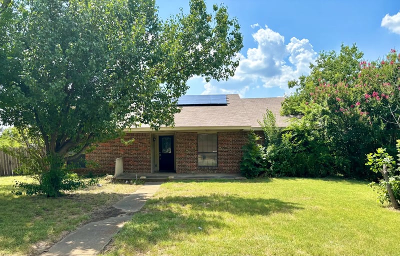 Single-story red brick home with rooftop solar panels, shaded by a large front-yard tree in Carrollton, Texas