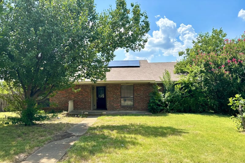 Single-story red brick home with rooftop solar panels, shaded by a large front-yard tree in Carrollton, Texas
