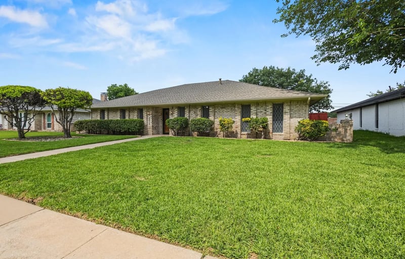Single-story tan brick ranch home with a low shingled roof and a manicured front lawn in Plano, Texas