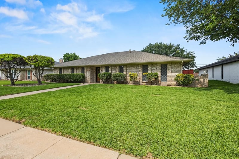 Single-story tan brick ranch home with a low shingled roof and a manicured front lawn in Plano, Texas