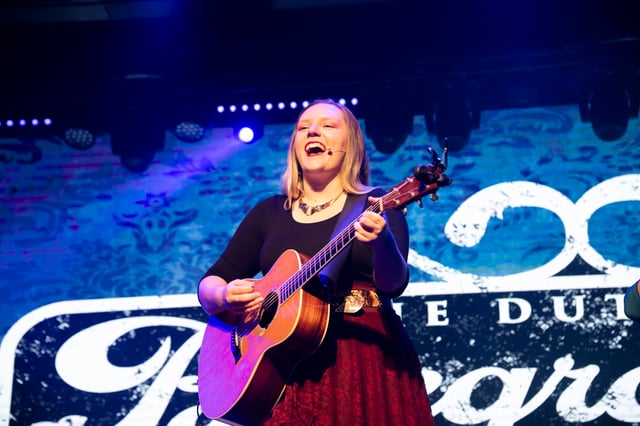 Jessica Dutton laughing as she plays acoustic guitar in front of the Bluegrassified sign