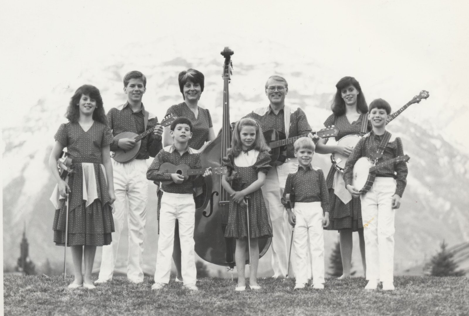 Vintage black-and-white photo of the young Dutton family holding fiddles, mandolins, banjos, guitars and an upright bass in front of snowy mountains