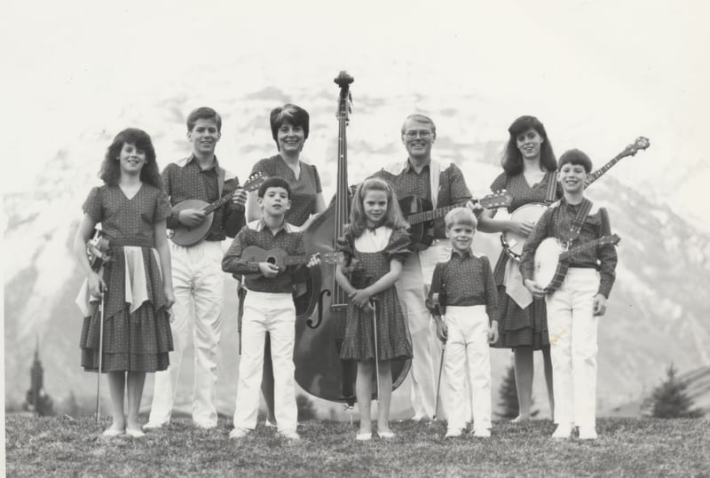 Vintage black-and-white photo of the young Dutton family holding fiddles, mandolins, banjos, guitars and an upright bass in front of snowy mountains