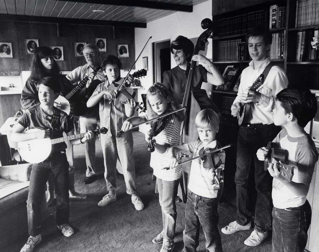 Vintage black-and-white photo of the Dutton children and parents playing fiddles, banjos, guitars and bass together in their living room