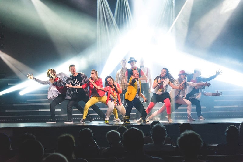 The Hughes Music Show cast singing and dancing under bright spotlights