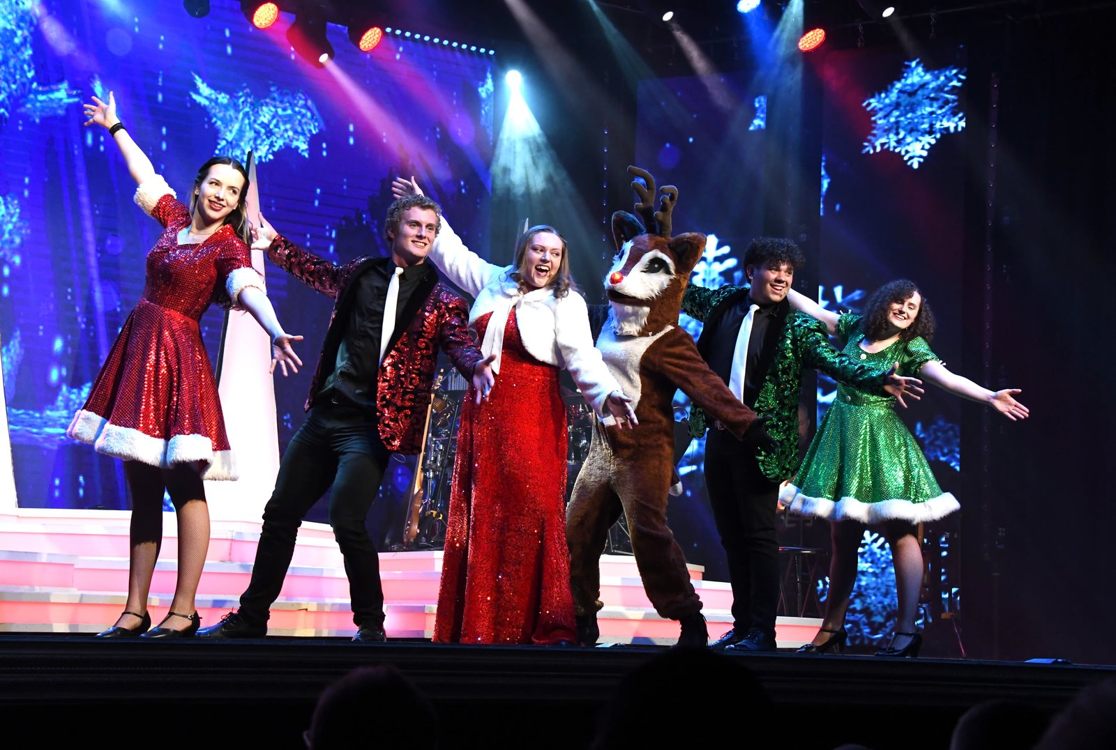 The Duttons cast in red and green Christmas costumes on stage with a dancing reindeer
