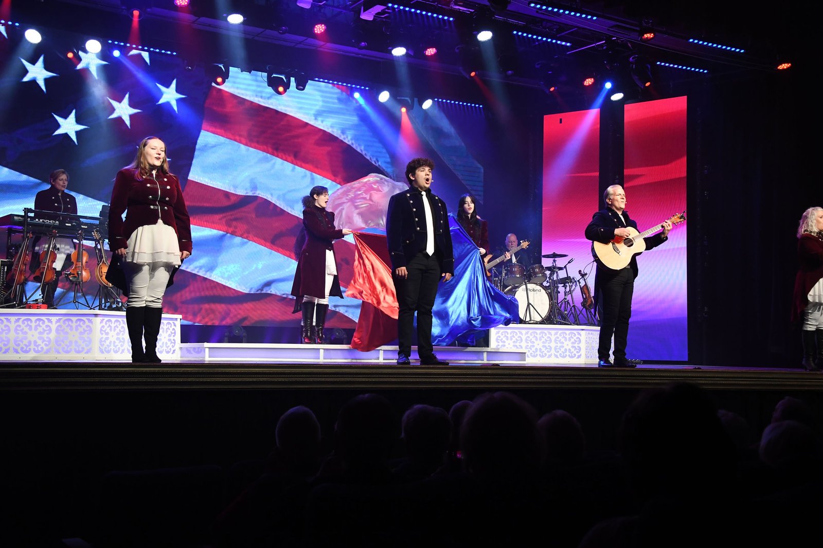 Singers in red and navy coats on stage in front of a giant waving American flag on the video wall