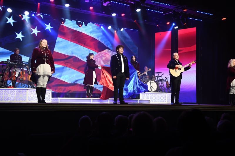 Singers in red and navy coats on stage in front of a giant waving American flag on the video wall
