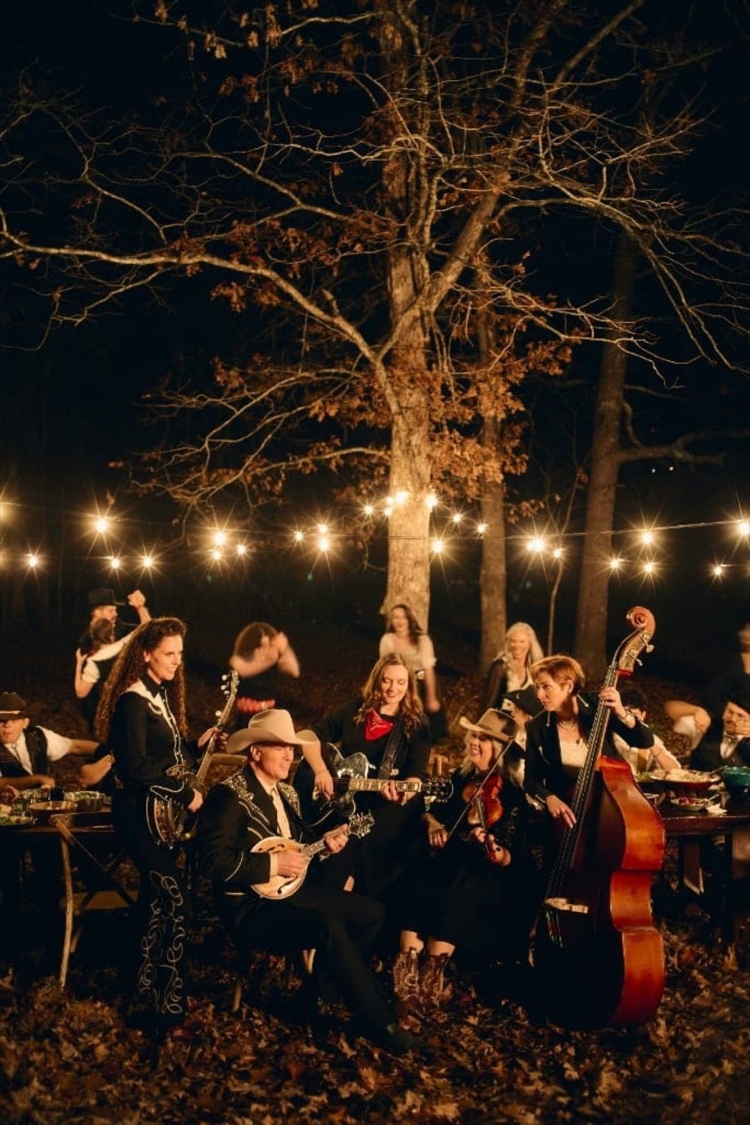 The Duttons playing banjo, guitars, fiddle, mandolin and upright bass under string lights at a nighttime outdoor gathering