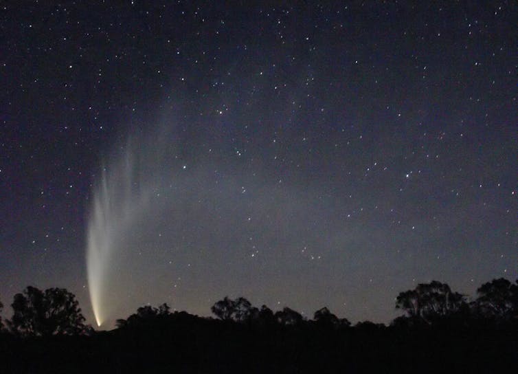A landscape with a bright arc of light across the night sky, coming out of a single point.