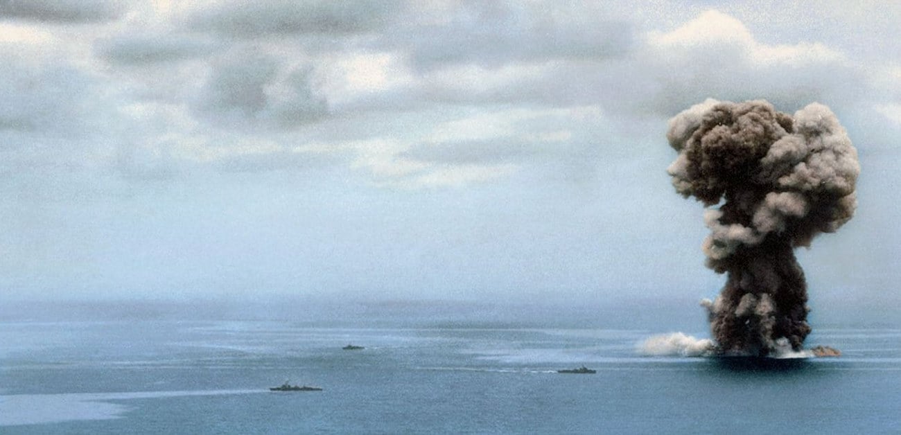 Explosion massive du Yamato, avec plusieurs navires visibles à distance. Un panache de fumée sombre et imposant s'élève de l'eau.