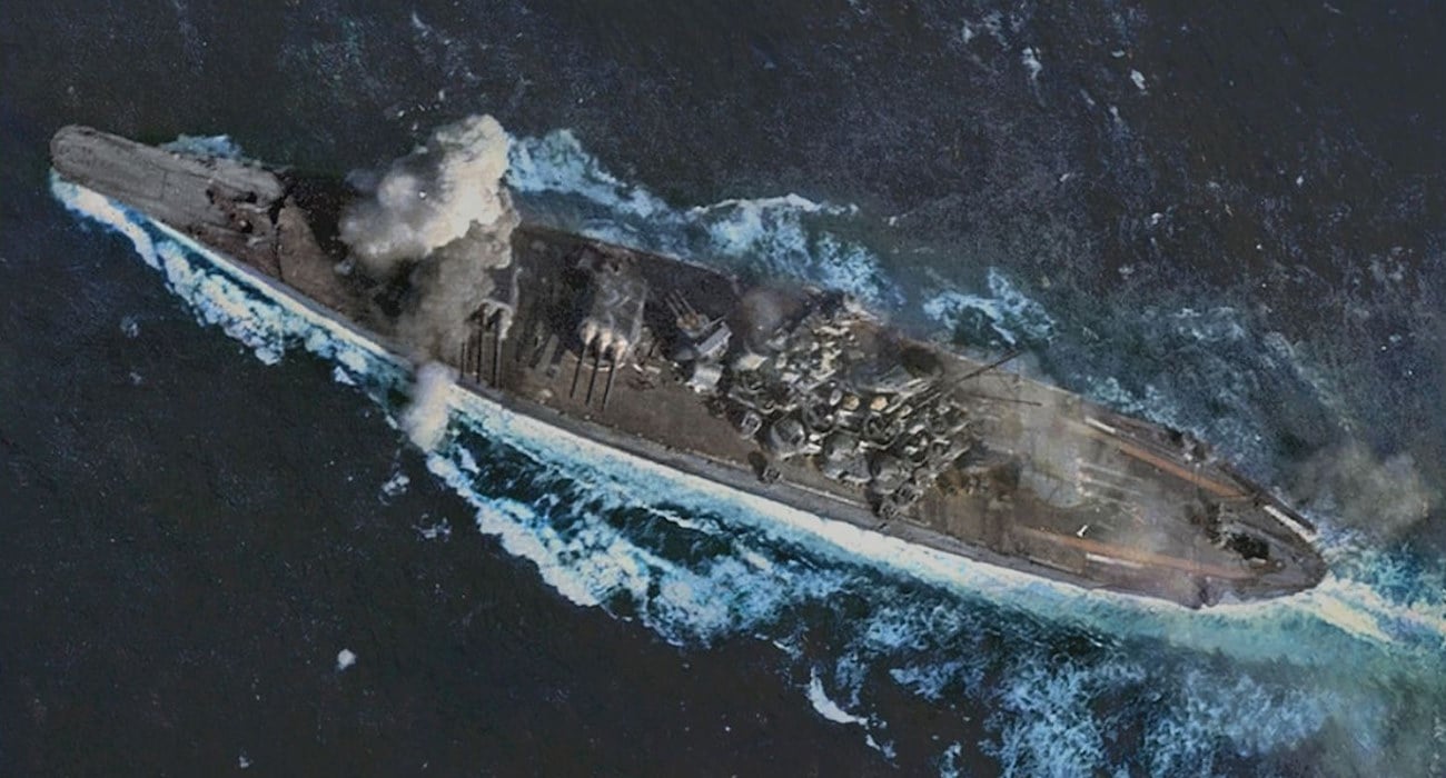 Vue aérienne du cuirassé japonais Yamato pendant la bataille du golfe de Leyte. Le navire subit des dégâts mineurs, visibles par quelques impacts et une légère fumée, mais reste globalement intact et poursuit sa navigation. La mer est agitée autour du bateau.