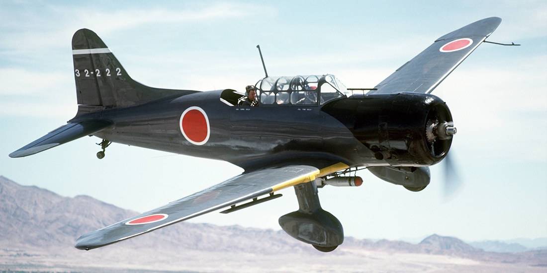 Avion bombardier en piqué japonais Aichi D3A Val en plein vol au-dessus d'un paysage montagneux, arborant les cocardes rouges japonaises (Hinomaru) sur un fuselage noir et gris.