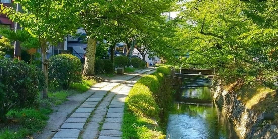 Chemin des philosophes à Kyoto en été avec canal d'eau, pavés de pierre et arbres verdoyants sous le soleil.