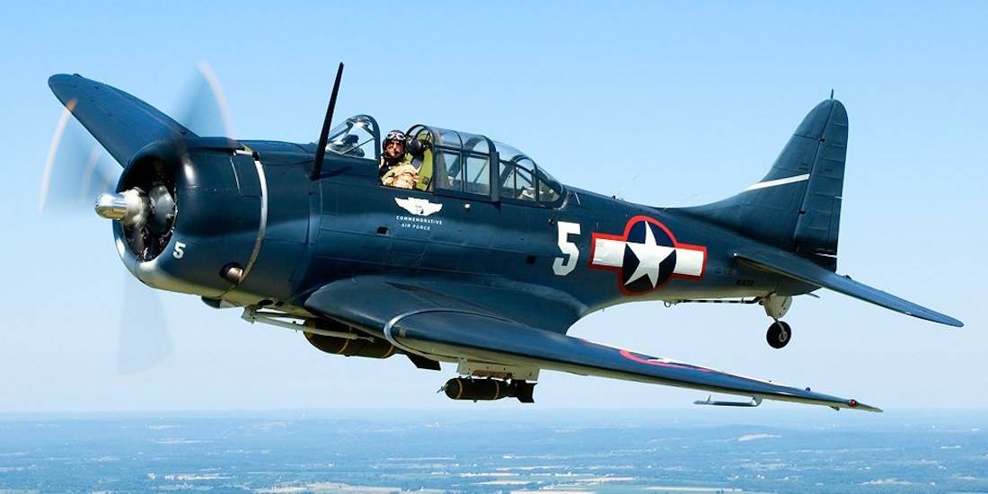 Bombardier en piqué Douglas SBD Dauntless de la Seconde Guerre mondiale en plein vol avec marquages de l'US Navy et pilote visible dans le cockpit.
