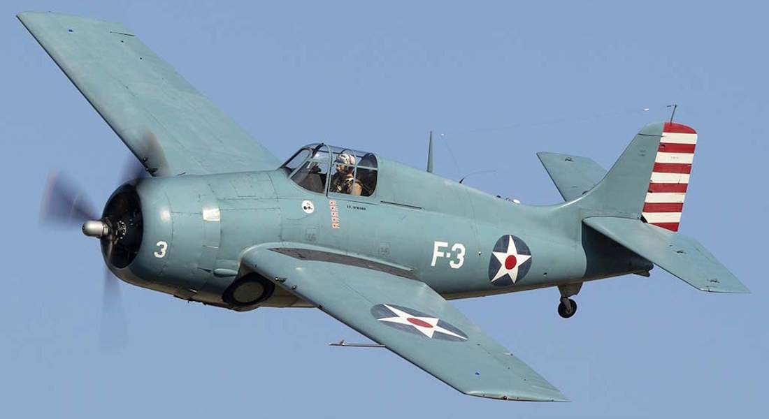 Avion de chasse américain Grumman F4F Wildcat en vol avec livrée historique de l'US Navy, cocardes à étoile rouge et dérive rayée sur fond de ciel bleu clair