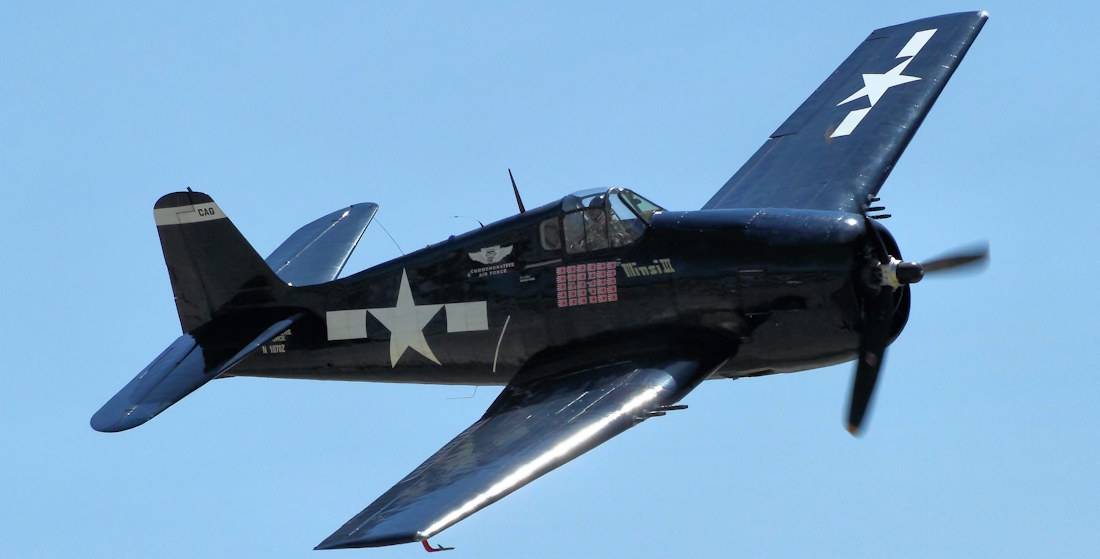 Avion de chasse Grumman F6F Hellcat bleu marine de la Seconde Guerre mondiale en plein vol sur fond de ciel bleu clair.