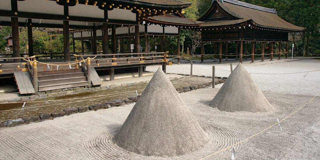 Cette photographie met en avant la cour intérieure du sanctuaire Kamigamo-jinja, classé au patrimoine mondial de l'UNESCO. Au premier plan, on aperçoit les deux monticules de sable purifié, ou Tatesuna, soigneusement façonnés pour représenter le mont Koyama. En arrière-plan, l'architecture classique de l'époque de Heian se dévoile avec ses structures en bois sombre et ses toits de chaume, illustrant parfaitement la sérénité et la tradition spirituelle japonaise au cœur de Kyoto. Deux cônes de sable rituels appelés Tatesuna devant les bâtiments en bois traditionnels du sanctuaire shinto Kamigamo-jinja à Kyoto, Japon.