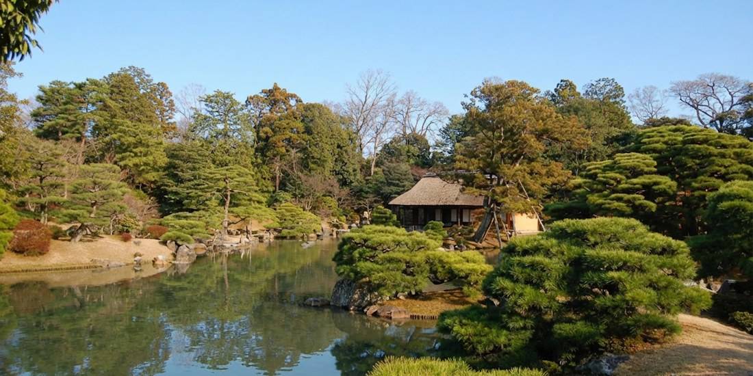 Vue panoramique du jardin japonais de la villa impériale de Katsura à Kyoto avec son étang central, ses pins taillés en nuages et un pavillon traditionnel au toit de chaume sous un ciel bleu clair.