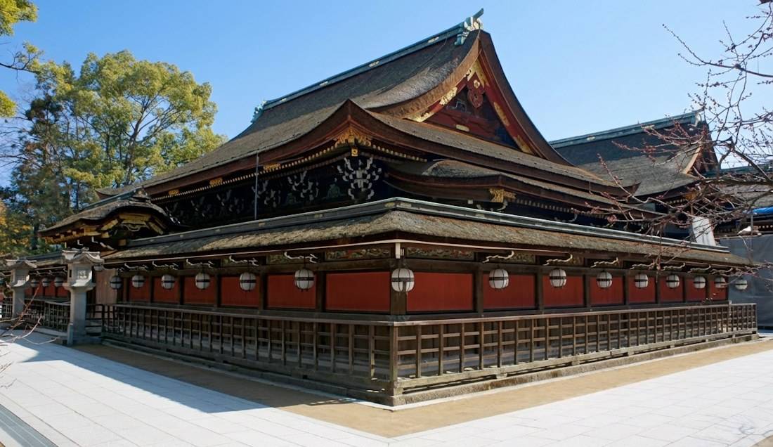 Vue extérieure du bâtiment principal en bois du sanctuaire Kitano Tenman-gu à Kyoto, Japon, montrant l'architecture traditionnelle shinto avec ses toits de chaume et ses lanternes blanches suspendues sous un ciel bleu.