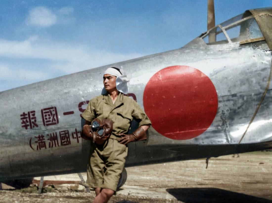 Pilote japonais en uniforme d'été posant devant un avion de chasse Mitsubishi A6M3 Zero avec l'insigne du soleil rouge (Hinomaru) sur le fuselage argenté.