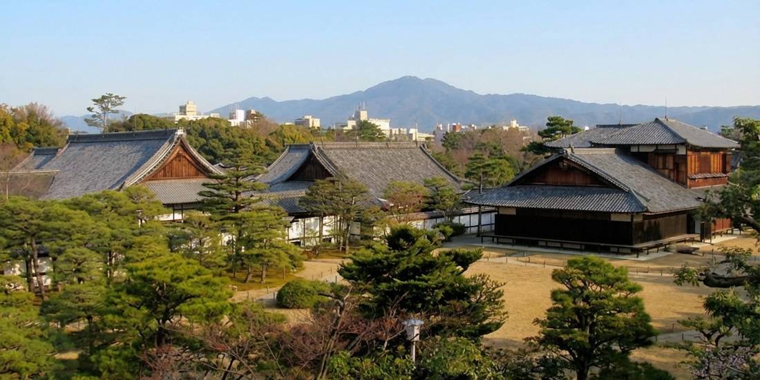 Vue panoramique du palais Ninomaru au château de Nijo à Kyoto, montrant l'architecture traditionnelle japonaise, les jardins zen et les montagnes en arrière-plan sous un ciel clair.