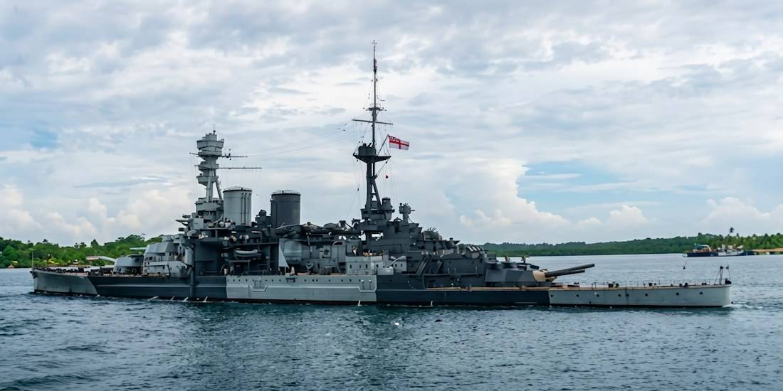 Croiseur de bataille britannique HMS Repulse quittant le port de Singapour en 1941 avec son camouflage de guerre gris et noir, naviguant sur une mer calme sous un ciel nuageux.