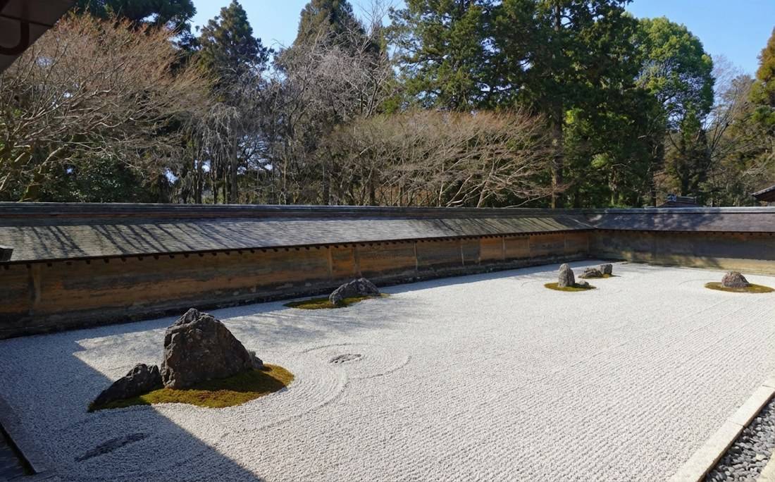 Jardin de pierre zen du temple Ryoan-ji à Kyoto avec gravier blanc ratissé et rochers entourés de mousse sous un ciel bleu.