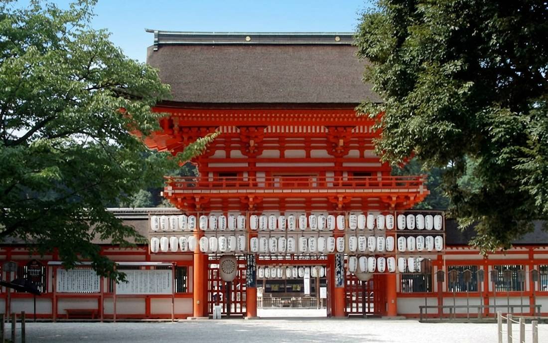 Vue de la porte monumentale vermillon (Romon) du sanctuaire Shimogamo-jinja à Kyoto, ornée de lanternes traditionnelles blanches sous un toit de chaume, entourée de verdure au Japon.