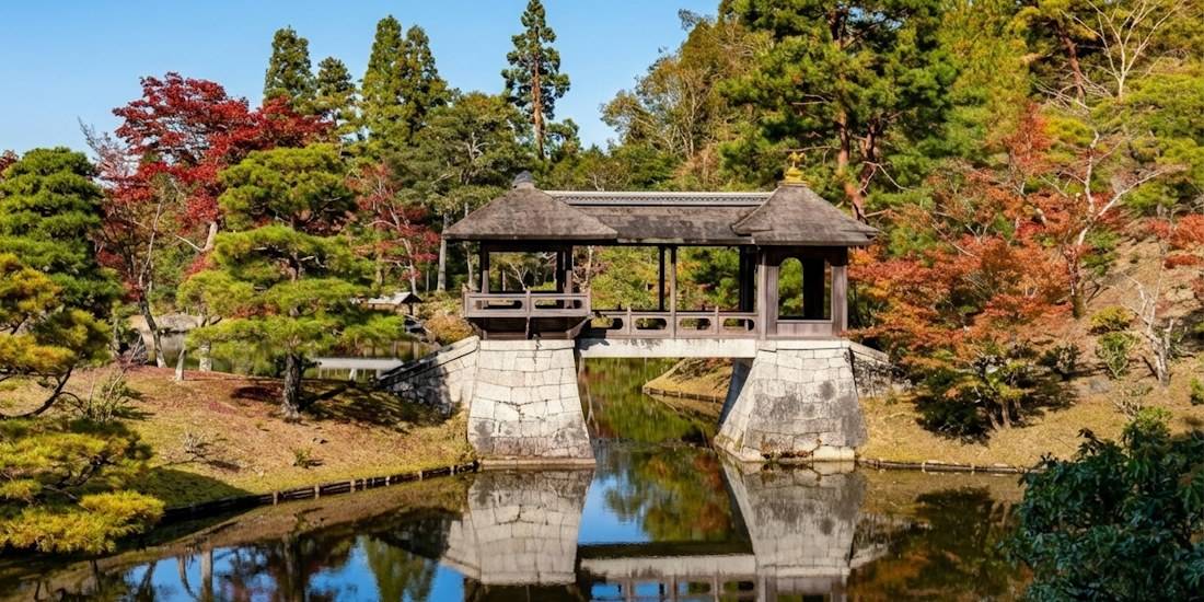 Pont couvert traditionnel en bois traversant un étang paisible à la villa impériale Shugaku-in de Kyoto, entouré de jardins japonais et de feuillages d'automne colorés.
