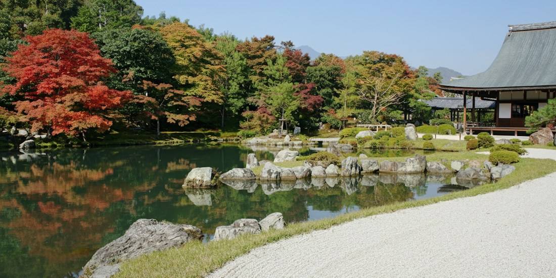 Vue panoramique du jardin zen Sogenchi au temple Tenryū-ji à Kyoto avec des érables aux couleurs d'automne se reflétant dans l'étang et un bâtiment traditionnel japonais.