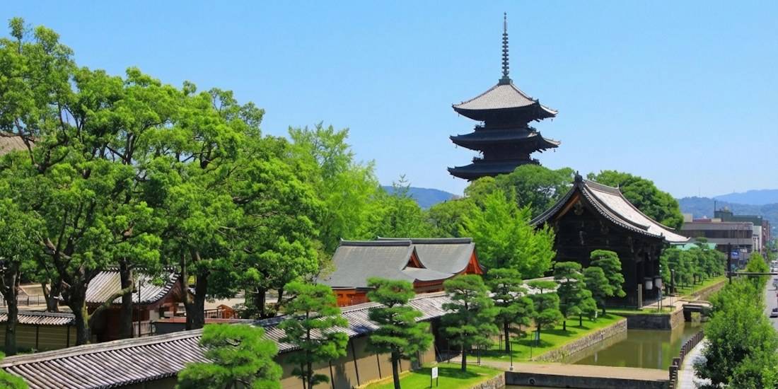 Vue panoramique du temple To-ji à Kyoto avec sa célèbre pagode à cinq étages s'élevant au-dessus des arbres verdoyants et des bâtiments traditionnels japonais sous un ciel bleu clair.