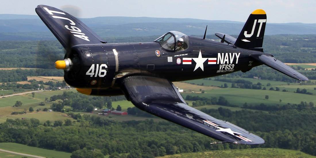 Avion de chasse Vought F4U Corsair bleu de l'US Navy en plein vol au-dessus d'un paysage rural verdoyant.