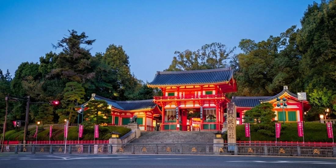 Vue de face du sanctuaire Yasaka-jinja à Kyoto au crépuscule, montrant la porte principale vermillon (Nishi-romon) illuminée avec des arbres verdoyants en arrière-plan sous un ciel bleu clair.