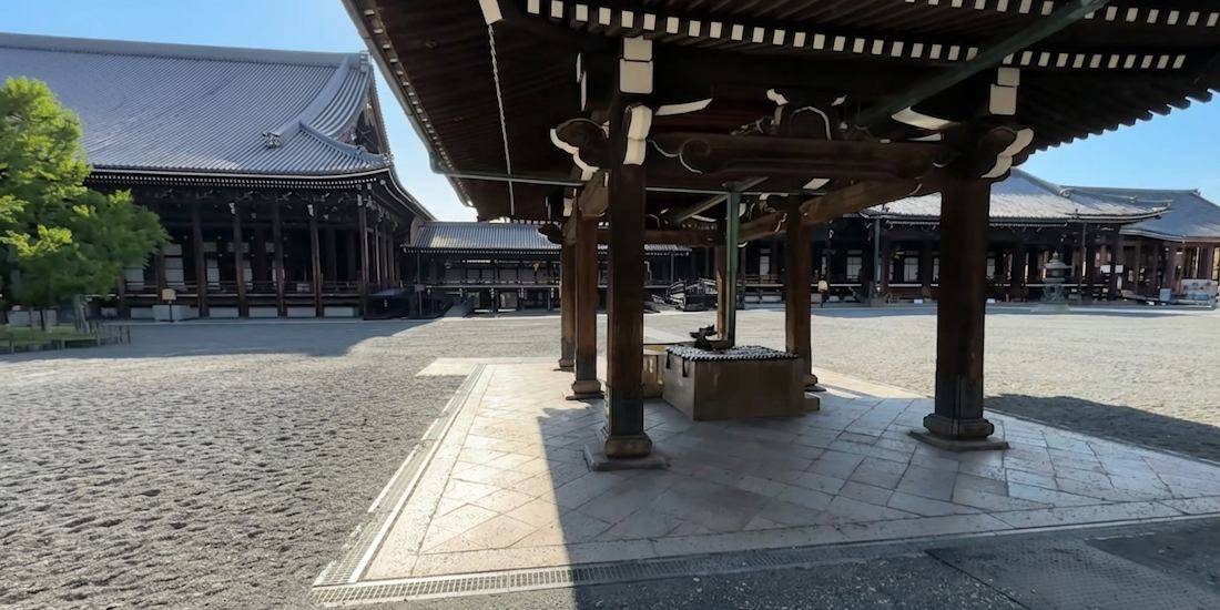 Chozuya traditionnel en bois au temple Nishi Hongan-ji à Kyoto, Japon, avec pavillons de temple bouddhiste en arrière-plan sous un ciel bleu.